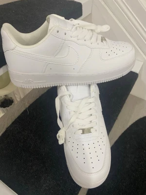 Nike Air Force 1 vita låga sneakers - Klassiska vita Nike Air Force 1 sneakers i lågt utförande. Skorna är helt vita med perforerad tå, vita skosnören och den ikoniska swoosh-loggan på sidan. Tillverkade i läder med en robust sula och diskreta detaljer i metall på snörningen.