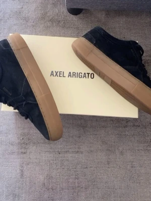 Svarta sneakers Axel Arigato mocka - Snygga svarta sneakers från Axel Arigato i mocka med bruna gummisulor. Klassisk låg modell med diskret logga på sidan och svarta skosnören. Perfekta för dig som gillar stilrena och trendiga skor.