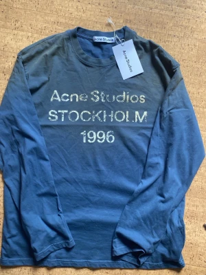 Mörkblå långärmad topp Acne Studios - Mörkblå långärmad topp från Acne Studios med tryck i vitt framtill där det står 'Acne Studios STOCKHOLM 1996'. Klassisk rund halsringning och avslappnad passform. Perfekt för dig som gillar stilrena och trendiga plagg. Använts otroligt mycket genom året, mycket vintage look. 