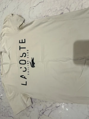 Vit Lacoste Sailing Club t-shirt - Snygg vit t-shirt från Lacoste med trycket 'Lacoste Sailing Club' och den klassiska krokodilen på bröstet. T-shirten har korta ärmar och är gjord i mjuk bomull för en skön känsla. Perfekt till jeans eller shorts för en clean och stilren look.
