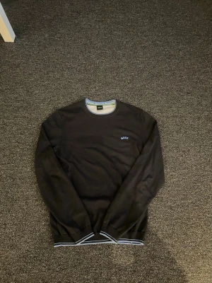 Svart tröja från BOSS med blå logga - Snygg svart tröja från BOSS med rund halsringning och diskret blå BOSS-logga på bröstet. Tröjan har ribbade muddar och gråvita detaljer vid hals, ärmslut och nederkant. Perfekt för en clean och stilren look.