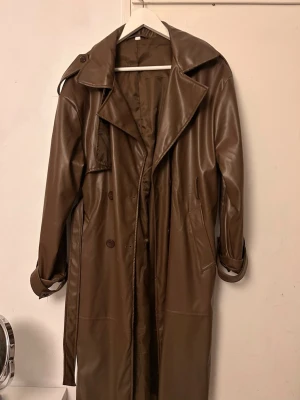 Brun trenchcoat i fejkläder - Snygg brun trenchcoat i fejkläder med klassisk dubbelknäppning, bred krage och bälte i midjan. Jackan har långa ärmar, axelklaffar och sidofickor. Perfekt för dig som vill ha en trendig och stilren look.