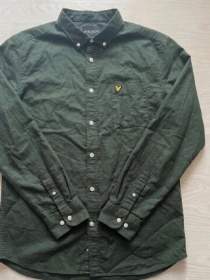 Grön skjorta från Lyle & Scott - Snygg mörkgrön skjorta från Lyle & Scott i 100% bomull. Klassisk button-down krage, vita knappar och ikonisk gul logga på bröstet. Perfekt till jeans eller chinos för en clean look.