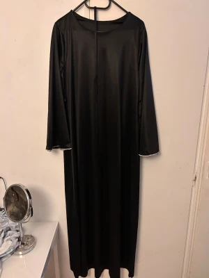 Svart långklänning med glittrig kant - En svart långklänning i glansigt material med långa ärmar och glittrig silverkant vid ärmsluten. Klänningen har en rak och enkel passform, perfekt för dig som gillar stilrena och eleganta plagg.