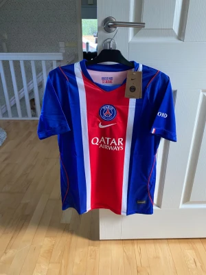PSG matchtröja blå/röd Nike - Snygg PSG matchtröja i blått, rött och vitt från Nike. Tröjan har klubbmärke och Qatar Airways-logga på bröstet, samt vita och röda detaljer längs framsidan. Tillverkad i lätt och ventilerande material, perfekt för fotboll eller träning.
