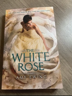 The White Rose av Amy Ewing - En ungdomsroman på engelska, 'The White Rose' av Amy Ewing. Omslaget har en elegant illustration av en ung kvinna i en ljus klänning, med titeln i blå text. Boken är en pocketutgåva från Harper Teen.