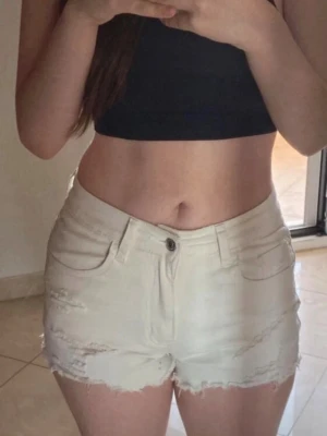 Vita högmidjade jeansshorts - Säljer ett par vita högmidjade jeansshorts med råa kanter och slitna detaljer. Shortsen har klassisk femficksdesign och knappgylf. Perfekta för varma dagar och ger en avslappnad look.