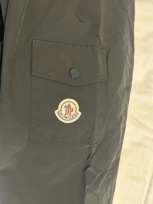 Svart vindjacka från Moncler - Snygg svart vindjacka från Moncler med klassisk logotyp på ärmen och praktisk ficka med knapp. Jackan har elastisk nederkant och innerfoder med serietidningsmönster. Tillverkad i lätt, glansigt material som ger en sportig look.