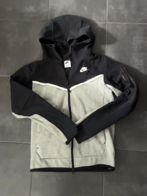 Nike Tech Fleece hoodie svart/grå - Nike Tech Fleece hoodie i svart och ljusgrått med dragkedja framtill, huva och fickor. Snygg vit Nike-logga på bröstet och en extra ficka med dragkedja på ärmen. Perfekt för en sportig och avslappnad stil.