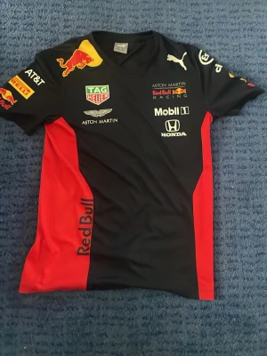 Red Bull Racing t-shirt från Puma - Snygg Red Bull Racing t-shirt från Puma i svart och rött med tryckta loggor från bland annat Aston Martin, Honda och Mobil 1. T-shirten har sportig design, rund hals och korta ärmar. Perfekt för motorsportfans som vill sticka ut.