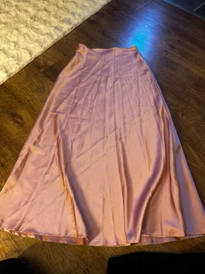 Ljusrosa långkjol i satin - Säljer en ljusrosa långkjol i mjukt satinliknande material. Kjolen har en enkel och stilren design med lätt glansig yta och faller snyggt längs benen. Perfekt att styla med en crop top eller stickad tröja för en trendig look. Använd en gång