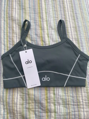 Mörkgrå sport-bh från Alo - Snygg mörkgrå sport-bh från Alo med tunna axelband och vita kontrastsömmar. Bh:n har en minimalistisk design med Alo-logga framtill och är tillverkad i ett stretchigt funktionsmaterial som passar perfekt till träning.