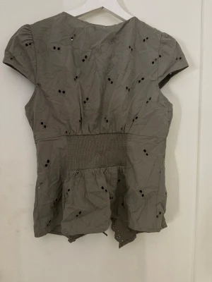 Olivgrön blus med broderade hål - Snygg olivgrön blus från GINA TRICOT med broderade hål och vågig spetskant framtill. Blusen har korta ärmar och smock i midjan för en skön passform. Perfekt för dig som gillar detaljer och en avslappnad men trendig stil.