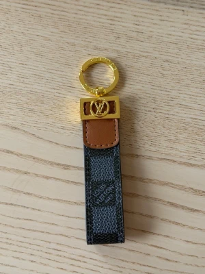  Vuitton nyckelring i canvas och läder - Snygg nyckelring från Vuitton i mörkgrå och svart rutmönstrad canvas med detaljer i brunt läder och guldfärgad metall. Klassisk LV-logga framtill och elegant design som passar perfekt till väskan eller nycklarna.