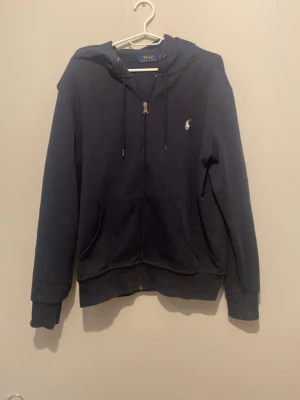 Mörkblå hoodie från Polo Ralph Lauren - Mörkblå hoodie med dragkedja från Polo Ralph Lauren. Klassisk design med huva, . Broderad logga i vitt på bröstet. Perfekt för en avslappnad och stilren look.