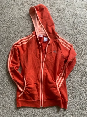 Röd Adidas hoodie med dragkedja - Snygg röd hoodie från Adidas med dragkedja och klassiska tre ränder längs ärmarna i ljusrosa. Hoodien har huva med ljusrosa insida och två fickor framtill. Perfekt för en sportig och avslappnad stil. Passar XS,S, 152