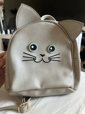 Ljusgrå kattryggsäck  - Supersöt liten ryggsäck i ljusgrått konstläder med kattansikte och utstickande kattöron. Väskan har justerbara axelband och dragkedja. Det ena bandet är pyttelite skrapat på sidan med det syns knappt