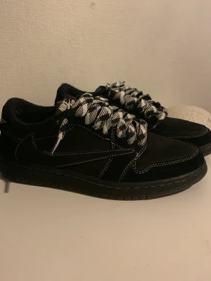 Travis scott black phantom  - Snygga svarta Nike sneakers i mocka med vita kontrastsömmar och tjocka svartvita mönstrade skosnören. Klassisk låg modell med diskret swoosh på sidan och perforerad tå för extra stil. Perfekta för dig som vill sticka ut med detaljer.