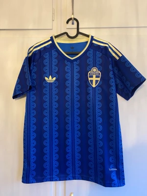 Blå Sverige fotbollströja Adidas - Snygg blå fotbollströja från Adidas med Sveriges landslagsemblem på bröstet och klassiska gula ränder på axlarna. Tröjan har ett unikt mönster i olika blå nyanser och v-ringad krage. Perfekt för match eller träning.