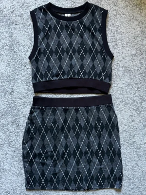 Stickad argyle topp från NLY Trend - Trendigt stickad topp från NLY Trend i storlek M. Ärmlös och croppad modell med mörkgrått, svart och vitt argylemönster. Ribbad kant vid hals och ärmhål för en clean finish. Perfekt för dig som gillar preppy vibes.