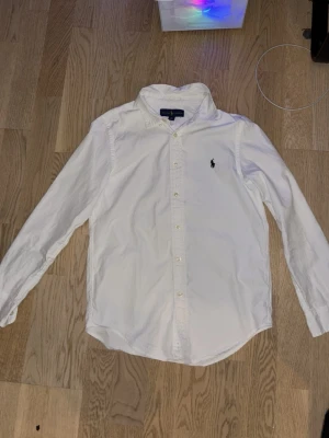 Vit skjorta från Ralph Lauren - Klassisk vit skjorta från Ralph Lauren med lång ärm och knappar framtill. Skjortan har den ikoniska broderade loggan på bröstet och är tillverkad i bomull. Perfekt för en clean och stilren look är 150 i storlek lite mindre än xs