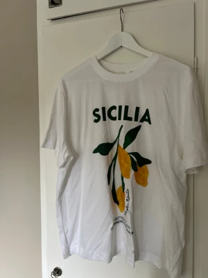 Vit t-shirt med citrontryck och text - Säljer en vit t-shirt med tryck av gula citroner och gröna blad samt texten 'SICILIA' i grönt på bröstet. T-shirten har rund hals och korta ärmar. Materialet känns som bomull och passformen är normal. Perfekt för dig som gillar fräscha och somriga prints.