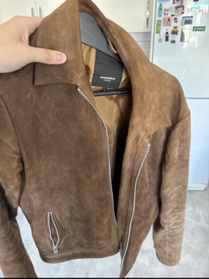 Brun mockajacka från RockAndBlue - Snygg och skön brun mockajacka från Roomarkable med silvriga dragkedjor och klassisk krage. Jackan har en stilren design och är tillverkad i mjukt skinn som ger en exklusiv känsla. Perfekt för dig som vill ha en tidlös och trendig look.