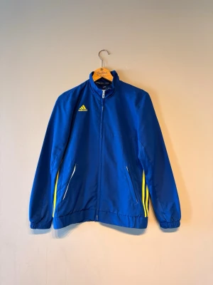 Blå träningsjacka från Adidas - Snygg blå träningsjacka från Adidas med dragkedja framtill och gula detaljer längs sidorna. Jackan har ståkrage, två fickor med dragkedja och Adidas-logga i gult på bröstet. Perfekt för träning eller sportiga dagar.