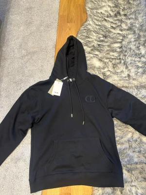 Svart hoodie med broderad logga från dior - Snygg svart hoodie med huva och dragsnören. På bröstet finns en diskret broderad logga med bokstäverna CD. Hoodien har en stor magficka och ribbade muddar vid ärmslut och nederkant. Perfekt för en chill och stilren look. Pris kan diskuteras
