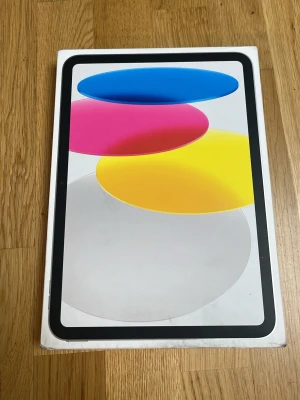 Apple iPad (A16) 128GB Wi-Fi - Säljer en Apple iPad (A16) med 128GB lagring och Wi-Fi. Surfplattan har en modern design med tunn ram och stor skärm. Färgglad förpackning med blå, rosa, gul och grå grafik. Perfekt för surf, spel och streaming.