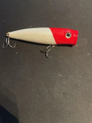 Röd och vit fiskedrag popper - Säljer ett fiskedrag i poppermodell med röd front och vit kropp. Draget har två trekrokar i metall och ett fisköga som detalj. 