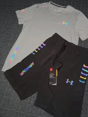 Grå träningsset från Under Armour - Säljer ett par grå träningsset från Under Armour med färgglada regnbågsdetaljer och logga på benet. Shortsen har snörning i midjan och tryckt text längs sidan. Perfekta för dig som vill sticka ut med sportig stil. Storlek S, helt ny 