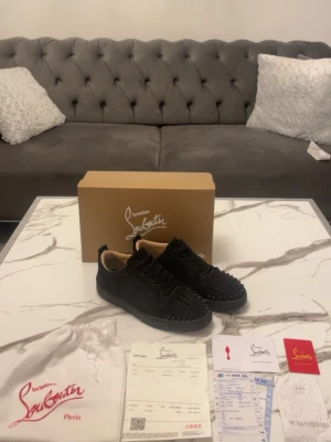 Svarta Louboutins  - Snygga svarta sneakers från Louboutin med coola svarta nitar över hela tån och sidorna. Skorna har en klassisk låg siluett, svart sula och är tillverkade i mocka och läder. Perfekta för dig som vill sticka ut med en edgy och exklusiv stil.