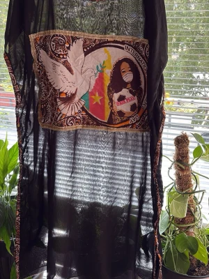 Svart transparent topp med tryck - Svart transparent topp med färgglatt tryck framtill som visar en kvinna, duva och flagga. Toppen har mönstrade kanter och är ärmlös med rund hals. Perfekt för dig som vill sticka ut med ett unikt plagg.