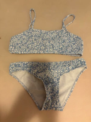 Blåvit bikini med blommönster - Söt bikini i blått och vitt med blommigt mönster. Överdelen har smala axelband och raka kanter, medan underdelen har klassisk brief-modell. Materialet är strukturerat och stretchigt för skön passform. Perfekt för strand eller poolhäng!