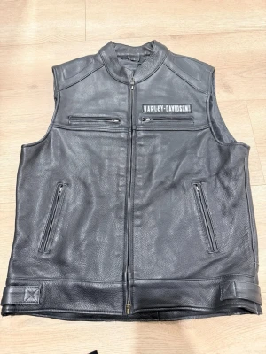 Svart Harley-Davidson läderväst - Cool svart läderväst från Harley-Davidson med dragkedja framtill och flera fickor med dragkedja. Stora broderade loggor i vitt och orange på ryggen samt mindre logga på bröstet. Perfekt för dig som gillar bikerstil och vill sticka ut.
