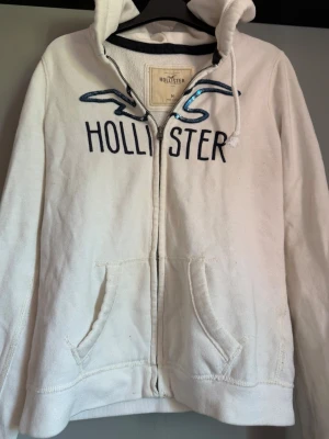 Vit hoodie med dragkedja från Hollister - Vit hoodie från Hollister med dragkedja framtill och två fickor. Stor logga och texten 'Hollister' i mörkblått på bröstet, samt blå paljettdetaljer. Mjuk bomullsmix och huva med dragsko. Perfekt för en chill och avslappnad stil. Priset går att diskutera!!