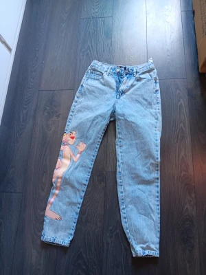 Ljusblå jeans med Pink Panther-tryck - Unika ljusblå mom jeans med ett stort tryck av Pink Panther på vänster ben. Klassisk femficksmodell med hög midja och raka ben. Tillverkade i slitstark denim, perfekta för dig som vill sticka ut med en cool och lekfull stil.
