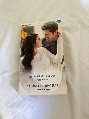 Ett mirakel till jul / Så länge hjärtat slår - En pocketbok med två romantiska berättelser: 'Ett mirakel till jul' av Kate Hardy och 'Så länge hjärtat slår' av Alison Roberts. Omslaget visar ett par i mysig vintermiljö framför en brasa. Perfekt för dig som gillar kärleksromaner med julkänsla.