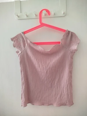 Ljusrosa off shoulder topp  - Säljer en ljusrosa off shoulder topp. Toppen har en ribbad, stretchig struktur och korta ärmar med volangkant. Perfekt för dig som gillar en feminin och trendig stil. Materialet känns mjukt och följsamt mot huden.