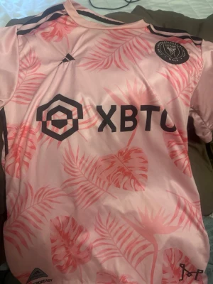 Rosa Inter Miami Messi matchtröja - Säljer en rosa Inter Miami matchtröja från Adidas med svarta detaljer och palmbladsmönster. Tröjan har tryck med Messi och nummer 10 på ryggen samt klubbmärke och sponsorlogga på framsidan. Tillverkad i ett lätt och ventilerande material.