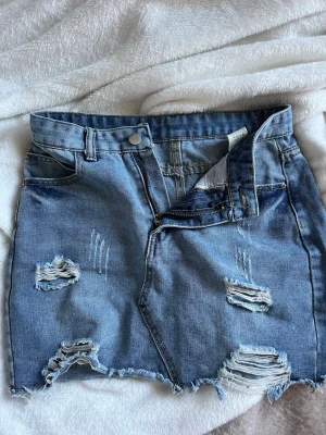 Blå jeanskjol med slitningar - Trendig blå jeanskjol i kort modell med råa kanter och slitna detaljer framtill. Vet tyvärr inte vart ifrån den är då jag ärvt den av min syster för ett tag sedan!💛Storlek S