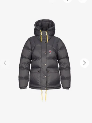 Grå dunjacka med huva från Fjällräven - Snygg grå dunjacka från Fjällräven med stor huva och gula dragskor. Jackan har två stora fickor framtill, tryckknappar och en liten Fjällräven-logga på bröstet. Perfekt för kalla vinterdagar och riktigt varm tack vare dunfyllningen.