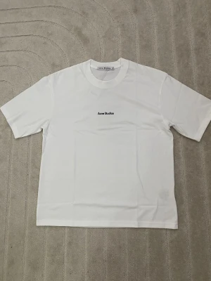 Vit Acne Studios t-shirt i bomull M - Klassisk vit t-shirt från Acne Studios med diskret logga tryckt på bröstet. T-shirten har rund halsringning och korta ärmar. Tillverkad i mjuk bomull för en clean och enkel look.