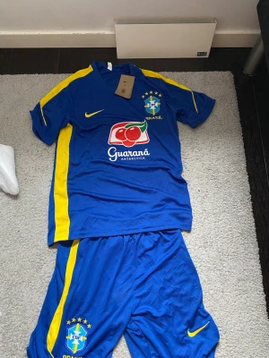 Blå Brasilien fotbollsställ Nike - Säljer ett blått Brasilien fotbollsställ från Nike med gula detaljer. Setet består av tröja och shorts, båda med CBF-logga och Brasil-tryck. Tröjan har Guaraná Antarctica sponsortryck och klassisk Nike swoosh. Perfekt för träning eller match! Pris kan diskuteras, andra frågor och funderingar svarar jag gärna på. Ha en bra dag !