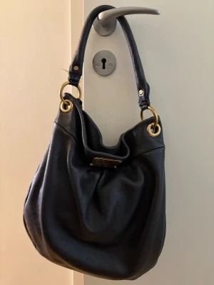 Svart läderväska Marc by Marc Jacobs - Snygg svart handväska i mjukt läder från Marc by Marc Jacobs. Väskan har en slouchy form, guldfärgade metalldetaljer och ett handtag med ringar. På framsidan sitter en metallbricka med märkets logga.