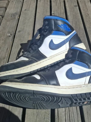 Svarta och blå Nike Air Jordan 1 High - Säljer ett par Nike Air Jordan 1 High sneakers i svart, vitt och blått. Skorna har klassisk hög siluett, svart läder på ovandelen, vita paneler och blå detaljer vid häl och Swoosh. Tjock vit sula och svart yttersula. Perfekta för dig som gillar streetwear och ikoniska sneakers.