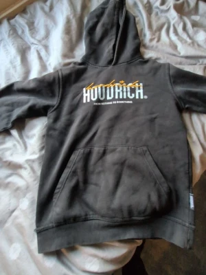 Svart hoodie från Hoodrich - Svart hoodie från Hoodrich med stor vit och gul logga på bröstet och texten 'FROM NOTHING TO SOMETHING'. Klassisk känguruficka framtill och huva. Perfekt för en streetwear-look och tillverkad i mjukt bomullsmaterial.