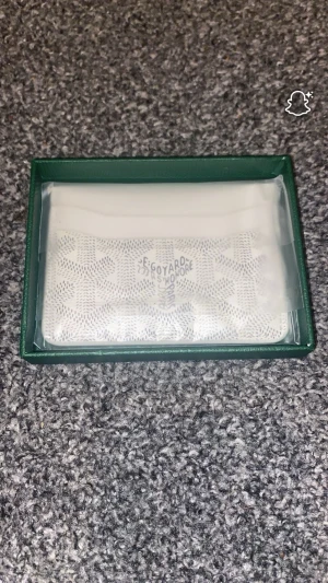 Vit plånbok i box - Säljer en stilren vit plånbok med klassiska mönster. Plånboken levereras i original grön box och har en minimalistisk design som passar perfekt för kort och sedlar. Materialet är troligtvis canvas med tryckt mönster.