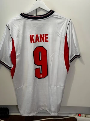 England fotbollströja Kane 9 Nike - Säljer en vit England fotbollströja med röda detaljer och svarta inslag. Tröjan har nummer 9 och namnet KANE på ryggen, samt Englands landslagsemblem och Nike-logga på bröstet. Perfekt för dig som vill stötta landslaget eller bära en snygg matchtröja.